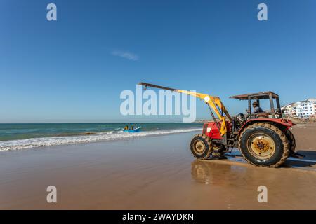 Un tracteur est sur la plage, prêt à soulever un petit bateau de pêche bleu en bois de l'eau après une sortie de pêche. Taghazout, Maroc, Afrique du Nord. Banque D'Images