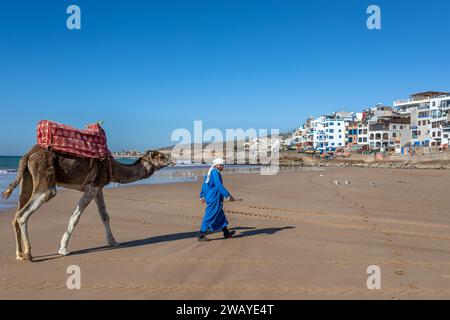 Homme berbère menant un chameau sur la plage de sable, avec des bâtiments et l'océan en arrière-plan Banque D'Images