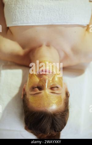 Le temps des soins de santé. Vue supérieure de la femme moderne détendue dans l'armoire de massage avec masque cosmétique doré sur le visage posé sur la table de massage. Banque D'Images