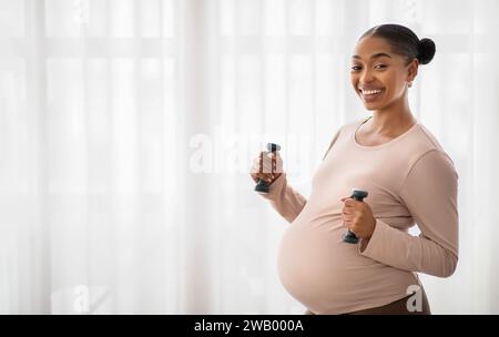 Femme noire enceinte souriante s'exerçant avec des haltères à la maison Banque D'Images