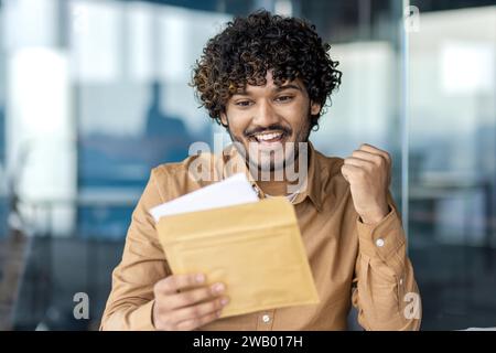 Homme d'affaires a reçu lettre de notification d'enveloppe postale avec de bonnes nouvelles, l'homme célèbre tenant la main vers le haut, lisant joyeusement les nouvelles, assis à l'intérieur du bureau avec ordinateur portable. Banque D'Images