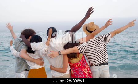 Groupe de jeunes amis divers se tenant dos à dos étreignant la mer tout en profitant de vacances Banque D'Images