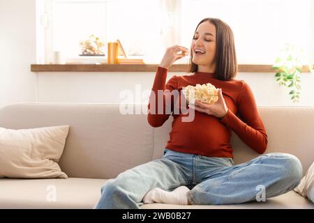 Joyeuse femme enceinte Eating Popcorn se relaxant sur le canapé à la maison Banque D'Images