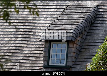 Un toit escarpé d'un bâtiment vintage avec des bardeaux en bois de cèdre. Il y a une longue lucarne recouverte de bois altéré. Le cadre de la fenêtre est de couleur verte. Banque D'Images