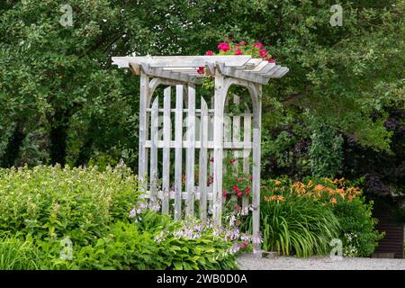 Une arche ou une tonnelle en bois en plein air entourée d'un jardin verdoyant et luxuriant. Le parc a des bouleaux, des roses rouges grimpantes, des fleurs de lis oranges Banque D'Images