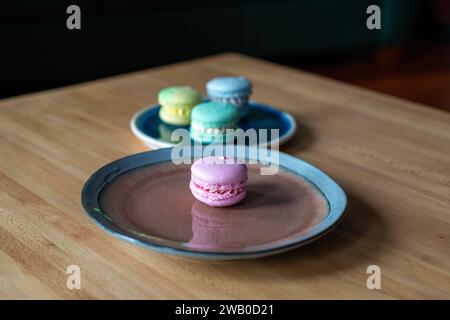 Quatre macarons français ; rose, vert, bleu et jaune. Les élégants desserts sucrés de style sandwich assortis sont sur une assiette bleue et brune et une table en bois. Banque D'Images