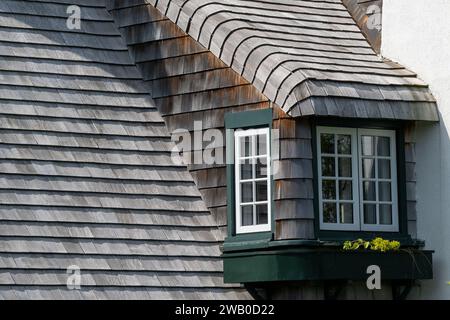 Un toit escarpé d'un bâtiment vintage avec des bardeaux en bois de cèdre. Il y a une longue lucarne recouverte de bois altéré. Le cadre de la fenêtre est peint en vert. Banque D'Images