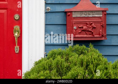 Une boîte aux lettres en métal rouge rétro vif, ou boîte aux lettres, est fixée au mur extérieur d'une maison en bois bleu avec une porte rouge vibrante. Banque D'Images