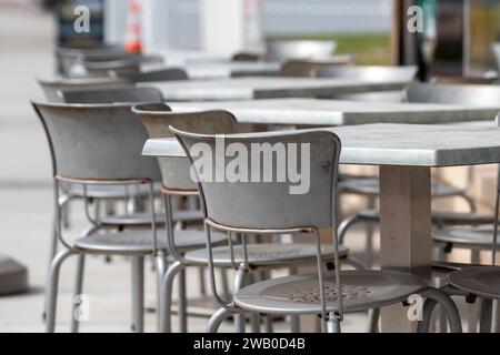 Une rangée de multiples tables carrées en métal gris vides et des chaises à l'extérieur sur le trottoir d'un café ou d'une boulangerie. Les chaises ont des dossiers ronds et sont vides. Th Banque D'Images