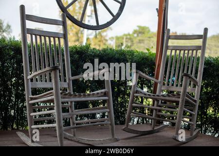 Deux chaises à bascule rustiques en bois, vieillies et usées, sur le porche d'une maison avec un arbuste vert luxuriant entourant la terrasse en bois. Banque D'Images