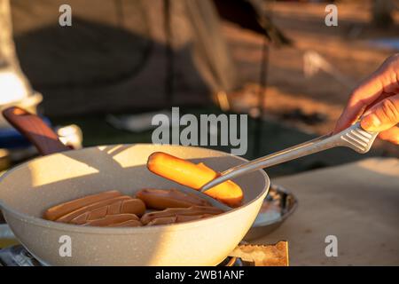 Une main de femme tient des saucisses grillées sur une poêle. Tout en allant camper pour se détendre Banque D'Images