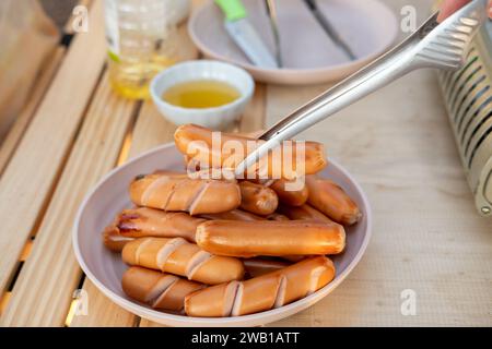 La main d'une femme tient des saucisses et les place sur une assiette. Tout en allant camper pour se détendre Banque D'Images