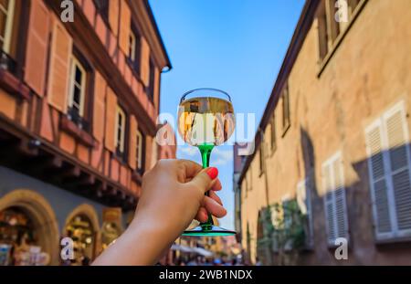 Main de femme avec un verre de vin Gewurztraminer dans un restaurant en plein air, maisons à colombages floues à Colmar France, route des vins d'Alsace village Banque D'Images