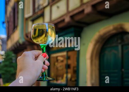 Main de femme avec un verre de vin Gewurztraminer dans un restaurant en plein air, maisons à colombages floues à Colmar France, route des vins d'Alsace village Banque D'Images