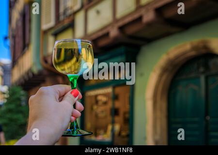 Main de femme avec un verre de vin Gewurztraminer dans un restaurant en plein air, maisons à colombages floues à Colmar France, route des vins d'Alsace village Banque D'Images