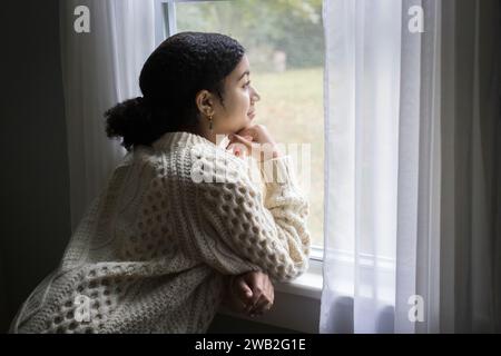 Biracial adolescente regarde par une fenêtre avec une expression paisible Banque D'Images