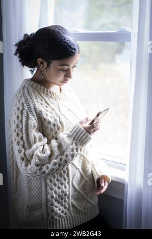 Biracial adolescente regarde son téléphone portable par la fenêtre Banque D'Images