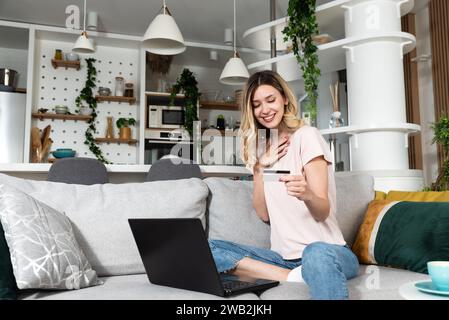 Jeune femme assise sur le canapé à l'aide d'un ordinateur portable et d'une carte de crédit pour les achats en ligne et commander des produits d'épicerie et de la nourriture pour la maison de sorte qu'elle n'a pas à le faire Banque D'Images