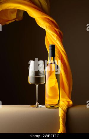 Bouteille et verre de vin blanc et volants de tissu jaune sur fond sombre. Banque D'Images