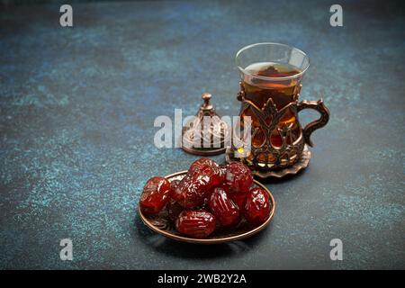 Rompre le jeûne avec des dattes séchées pendant le Ramadan Kareem, repas d'iftar avec des dattes et du thé arabe en verre traditionnel, vue d'angle sur fond bleu rustique Banque D'Images
