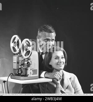 Projecteur de film dans le couple de 1960ez à un projecteur de film qui fonctionne. Ils ont les yeux sur l'écran où le film est projeté. Le film était généralement montré sur un écran blanc. Le rouleau avant du projecteur contient le film prêt à être vu, le rouleau arrière est le film enroulé au fur et à mesure qu'il est vu. Une fois le film terminé, la bobine arrière contenant le film a été enroulée sur la bobine d'origine. Suède 1965. Kristoffersson réf DG48-9 Banque D'Images