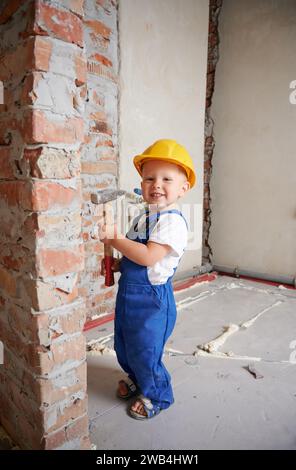 Toute la longueur de travailleur de construction d'enfant heureux tenant l'outil de marteau et regardant la caméra tout en se tenant par le mur de brique à la maison pendant la rénovation. Enfant souriant portant un casque de sécurité et une combinaison de travail. Banque D'Images