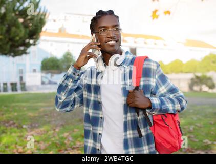 Joyeux jeune étudiant noir parlant au téléphone marchant à l'extérieur Banque D'Images