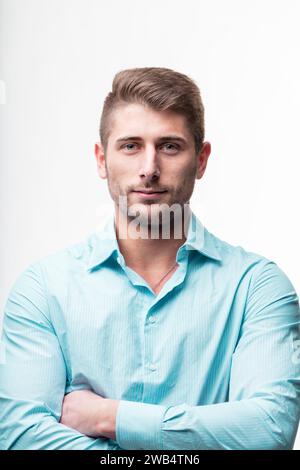 Jeune homme au regard confiant, bras croisés, portant une chemise rayée bleu clair sur fond blanc Banque D'Images