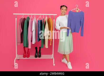 Femme asiatique joyeuse à côté du rack de mode démontrant de nouveaux vêtements Banque D'Images