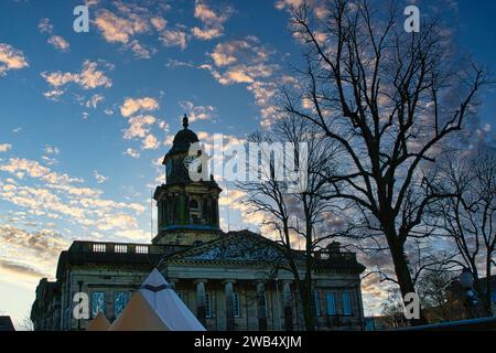 Bâtiment historique avec tour d'horloge au crépuscule, silhouettes d'arbres au premier plan, nuages roses dans le ciel bleu à Lancaster. Banque D'Images