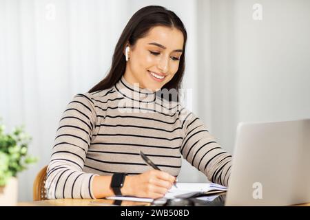 Jeune femme concentrée avec des écouteurs sans fil travaille assidûment à son ordinateur portable, prenant des notes Banque D'Images