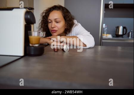 Belle brune aux cheveux bouclés endormie, jeune adulte jolie femme en peignoir blanc, appuyée sur le comptoir de cuisine et admirant le café capsule mac Banque D'Images