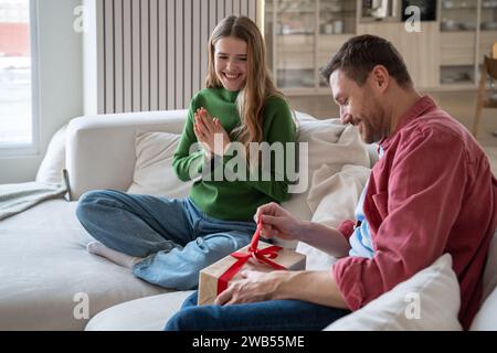 Joyeuses vacances en famille. Femme joyeuse regardant des montres aimant mari heureux ouvert surprise dans la boîte cadeau. Banque D'Images