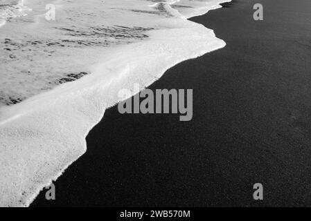 Plage de sable noir principalement floue avec mousse blanche de fond de vagues de mer Banque D'Images