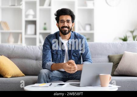 Homme indien souriant portant des lunettes assis à table avec ordinateur portable à la maison Banque D'Images
