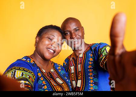 Homme noir souriant et femme portant des vêtements ethniques prenant selfie sur smartphone et regardant la caméra avant. Joyeux couple tenant le téléphone portable et faisant le portrait pov photo Banque D'Images