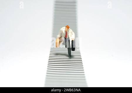 Photographie miniature de figurine de jouet de personnes minuscules. Un homme portant un manteau blanc sur un passage de zèbre à la rue. Isolé sur fond blanc. Image photo Banque D'Images