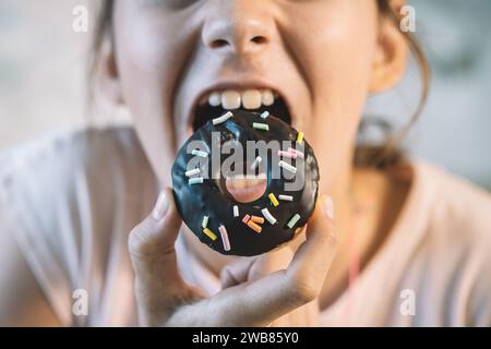 Petite fille mangeant un beignet messily. Un cupcake sucré à la main. La fille mange des pâtisseries sucrées. une bouche ouverte et un beignet au chocolat. Banque D'Images