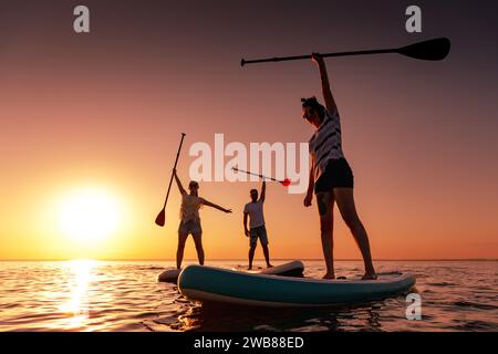 Trois heureux jeunes amis marchent sur des planches de sup ou paddleboards au coucher du soleil lac calme Banque D'Images