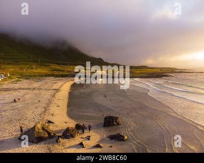 Vue aérienne sur la plage de Skagsanden près de Lofoten Beach Camp sous le soleil de minuit, le soleil et les nuages de basse montagne. Touristes sur la plage. Banque D'Images