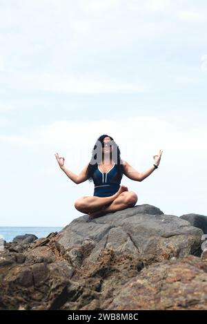 Belle femme aux cheveux bouclés assise sur le rocher d'une plage faisant des exercices de yoga. Personne heureuse voyageant. Banque D'Images