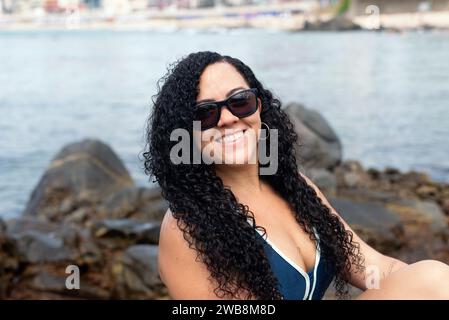 Belle femme aux cheveux bouclés portant des lunettes de soleil regardant la caméra contre la mer en arrière-plan. Personne heureuse voyageant. Banque D'Images