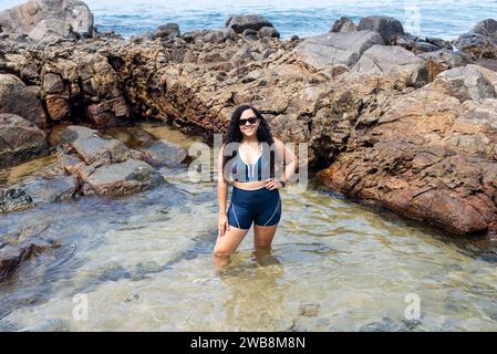 Belle femme aux cheveux bouclés portant des lunettes de soleil debout dans une flaque d'eau sur la plage. Personne voyageant. Banque D'Images