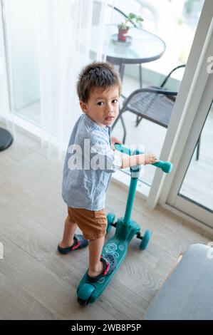 Découvrez la joie pure d'un garçon de deux ans et demi, vêtu d'une chemise et d'un bermuda, alors qu'il explore la pièce de manière ludique sur son baby Push sc Banque D'Images