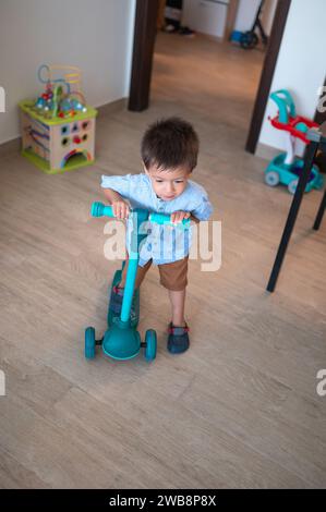 Découvrez la joie pure d'un garçon de deux ans et demi, vêtu d'une chemise et d'un bermuda, alors qu'il explore la pièce de manière ludique sur son baby Push sc Banque D'Images