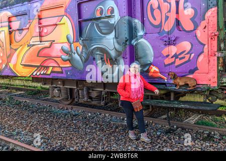 AS, Limbourg, Belgique. 17 décembre 2023. Touriste senior posant avec son teckel à côté d'un wagon peint avec des graffitis, pistes désaffectées à Banque D'Images