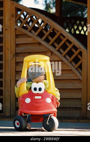 Portrait de mignon petit garçon caucasain en bas âge profiter d'avoir du plaisir à cheval voiture jouet dans le parc de la ville au jour du printemps. Banque D'Images