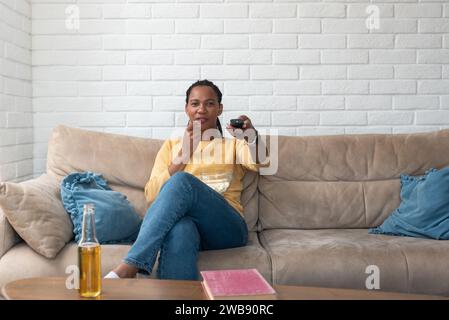 Rire jeune jolie femme afro-américaine assise sur le canapé, mangeant du pop-corn et regardant la télévision à la maison, tenant la télécommande, profitant d'un beau film A. Banque D'Images