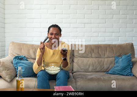 Rire jeune jolie femme afro-américaine assise sur le canapé, mangeant du pop-corn et regardant la télévision à la maison, tenant la télécommande, profitant d'un beau film A. Banque D'Images