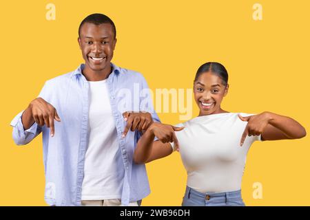 Joyeux couple noir pointant vers le bas, sourires fiers, fond jaune Banque D'Images